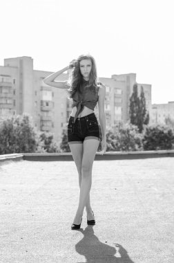 Mavi gökyüzüne ve modern evlere karşı çatıda oturan esmer Asyalı kız. Uçan saçlı genç bir kadın günlük kıyafetler giyer. Kot şort siyah gömlek giyer.