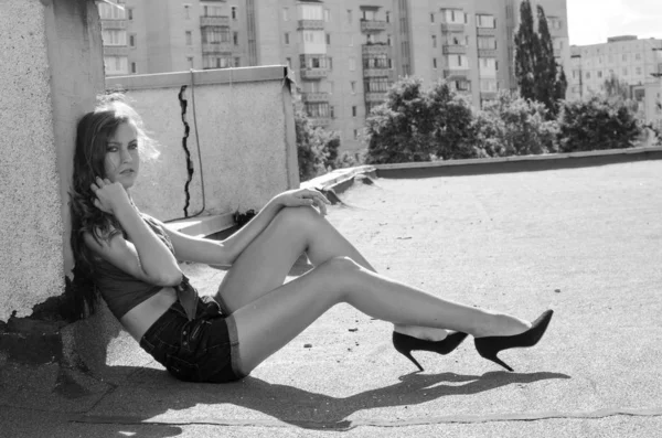 Assis brunette asiatique fille sur le toit contre le ciel bleu et les maisons modernes. Jeune femme aux cheveux volants porter une robe décontractée - jeans shorts chemise noire — Photo