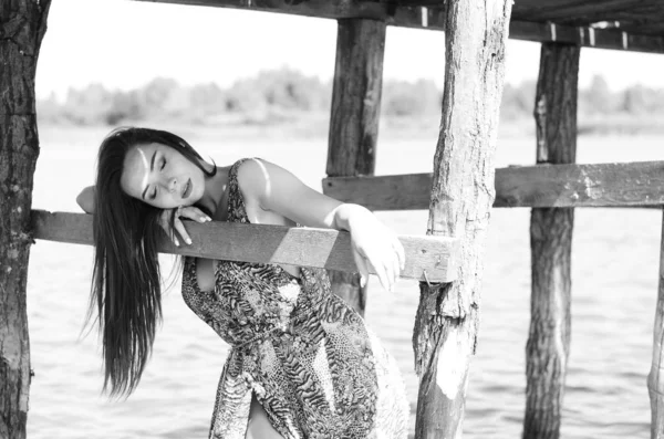 Mujer morena posando en el muelle de madera del lago. Mujer en traje de verano con la espalda vuelta a la pasarela de madera del lago Neusiedl. Reflejos solares en la casa de botes en el fondo . — Foto de Stock