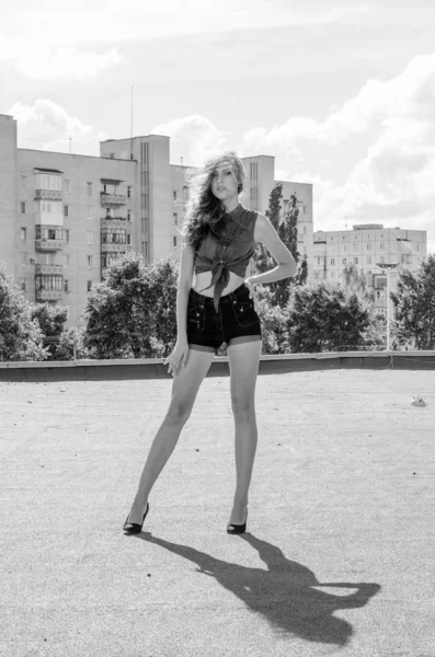 Sitting brunette asian girl on the roof against blue sky and modern houses. Young woman with flying hairs wear casual dress - jeans shorts  black shirt — Stock Photo, Image