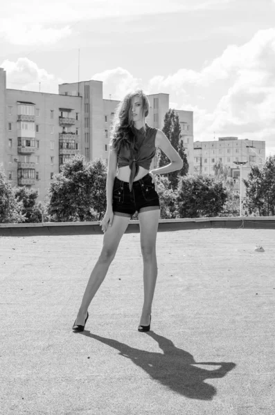 Sitting brunette asian girl on the roof against blue sky and modern houses. Young woman with flying hairs wear casual dress - jeans shorts  black shirt — Stock Photo, Image