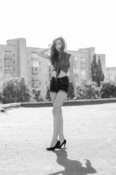 Sittande brunett asiatisk flicka på taket mot blå himmel och moderna hus. ung kvinna med flygande hår bära casual klänning - jeans shorts svart skjorta — Stockfoto