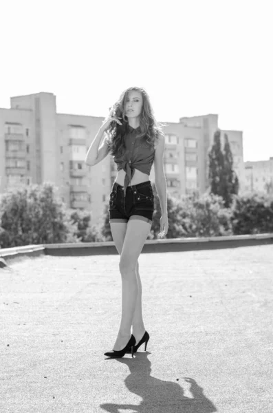 Sitting brunette asian girl on the roof against blue sky and modern houses. Young woman with flying hairs wear casual dress - jeans shorts  black shirt — Stock Photo, Image