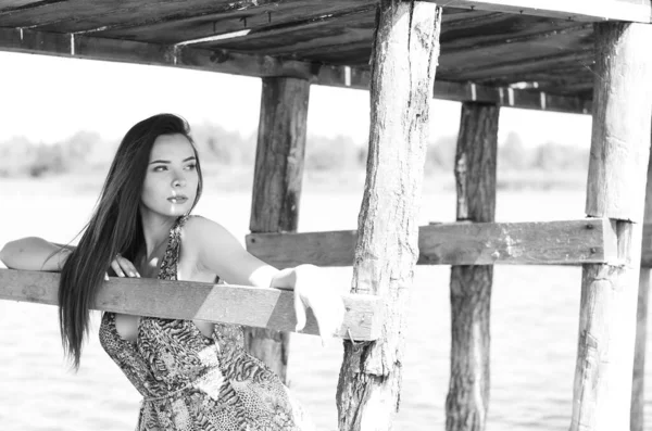 Mujer morena posando en el muelle de madera del lago. Mujer en traje de verano con la espalda vuelta a la pasarela de madera del lago Neusiedl. Reflejos solares en la casa de botes en el fondo . — Foto de Stock