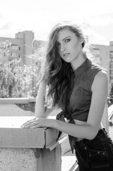 Sitting brunette asian girl on the roof against blue sky and modern houses. Young woman with flying hairs wear casual dress - jeans shorts  black shirt — Stock Photo, Image