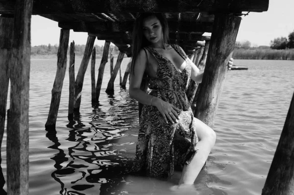 Mulher loira atraente sentada no cais ao lado do lago com fundo da natureza. noiva romântica vestindo vestido branco e segurando buquê de flores — Fotografia de Stock