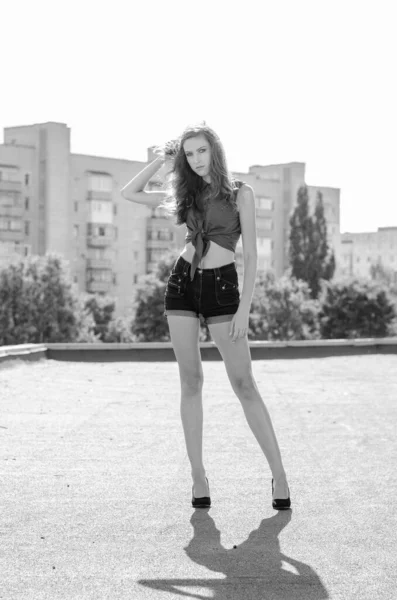 Zittend brunette aziatisch meisje op het dak tegen de blauwe lucht en moderne huizen. Jonge vrouw met vliegende haren dragen casual jurk - jeans shorts zwart shirt — Stockfoto