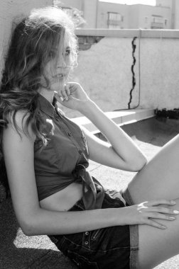 Mavi gökyüzüne ve modern evlere karşı çatıda oturan esmer Asyalı kız. Uçan saçlı genç bir kadın günlük kıyafetler giyer. Kot şort siyah gömlek giyer.