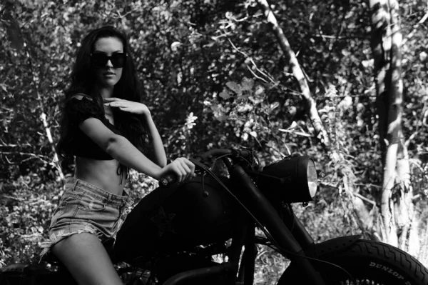 Junge sexy Frau mit langen welligen Haaren in Lederkleidung und Sonnenbrille posiert neben einem Motorrad. Schwarz-Weiß-Foto. — Stockfoto