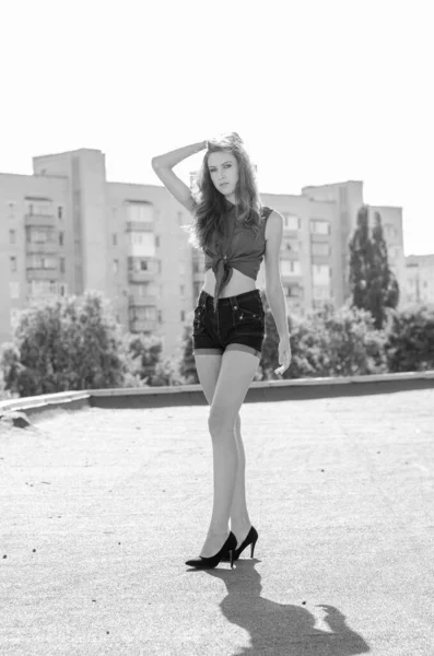 Sittande brunett asiatisk flicka på taket mot blå himmel och moderna hus. ung kvinna med flygande hår bära casual klänning - jeans shorts svart skjorta — Stockfoto