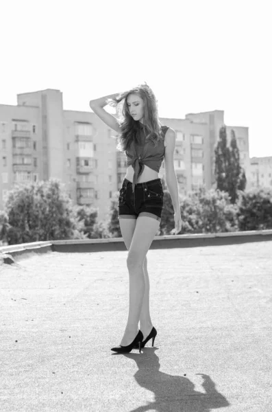Zittend brunette aziatisch meisje op het dak tegen de blauwe lucht en moderne huizen. Jonge vrouw met vliegende haren dragen casual jurk - jeans shorts zwart shirt — Stockfoto