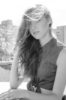 Mavi gökyüzüne ve modern evlere karşı çatıda oturan esmer Asyalı kız. Uçan saçlı genç bir kadın günlük kıyafetler giyer. Kot şort siyah gömlek giyer.