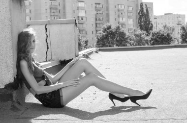 Mavi gökyüzüne ve modern evlere karşı çatıda oturan esmer Asyalı kız. Uçan saçlı genç bir kadın günlük kıyafetler giyer. Kot şort siyah gömlek giyer.