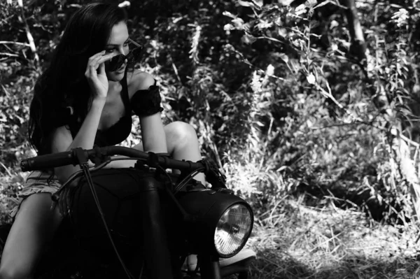 Jeune femme sexy avec de longs cheveux ondulés dans des vêtements en cuir et des lunettes de soleil posant près d'une moto. Photo noir et blanc. — Photo