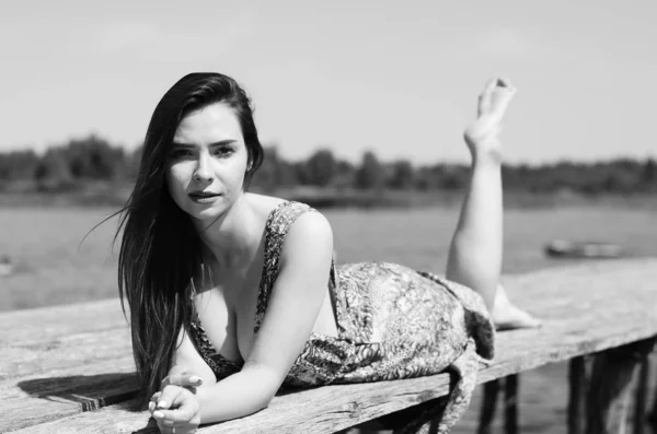 Donkerharige vrouw poseert bij de houten pier bij het meer. Vrouw in zomerkleding met haar rug naar de houten voetgangersbrug van het Neusiedlmeer. Zon reflecties bij het boothuis op de achtergrond. — Stockfoto