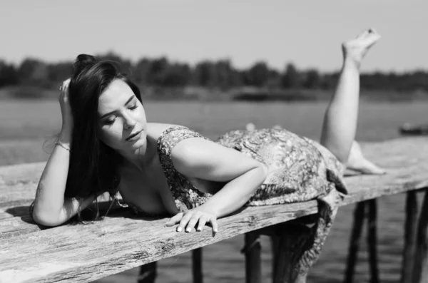 Sötét hajú nő pózol a fából készült mólónál a tónál. A nyári ruhás nő hátat fordított a Fertő-tó fából készült hídjának. Nap tükröződések a csónakházban a háttérben. — Stock Fotó