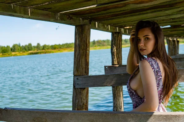 Mujer morena posando en el muelle de madera del lago. Mujer en traje de verano con la espalda vuelta a la pasarela de madera del lago Neusiedl. Reflejos solares en la casa de botes en el fondo . — Foto de Stock