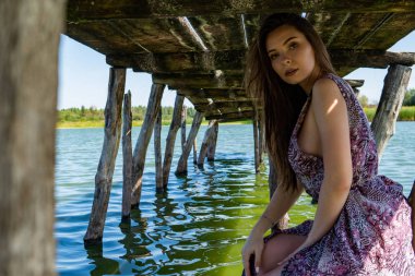 Gölün oradaki tahta iskelede poz veren siyah saçlı kadın. Sırtı Neusiedl Gölü 'nün tahta yaya köprüsüne dönmüş bir kadın. Arka plandaki kayıkhanede güneş yansımaları.