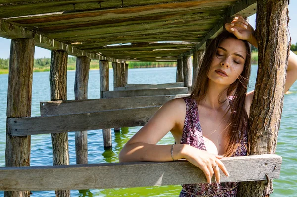 Mulher de cabelos escuros posando no cais de madeira no lago. Mulher em roupa de verão com as costas viradas para a passarela de madeira do Lago Neusiedl. Reflexões do sol na casa do barco no fundo . — Fotografia de Stock