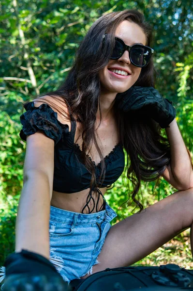 Mujer sexy joven con el pelo ondulado largo en ropa de cuero y gafas de sol posando cerca de una motocicleta. Foto en blanco y negro . — Foto de Stock