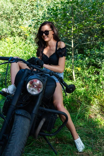 Młoda seksowna kobieta z długimi falowanymi włosami w skórzanych ubraniach i okularach przeciwsłonecznych, pozująca w pobliżu motocykla. Czarno-białe zdjęcie. — Zdjęcie stockowe
