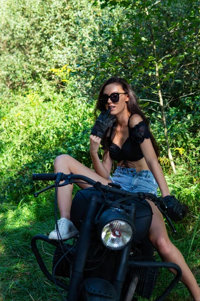 年轻性感的女人，留着长长的波浪般的头发，穿着皮衣，戴着太阳镜，站在摩托车旁边。黑白照片. — 图库照片