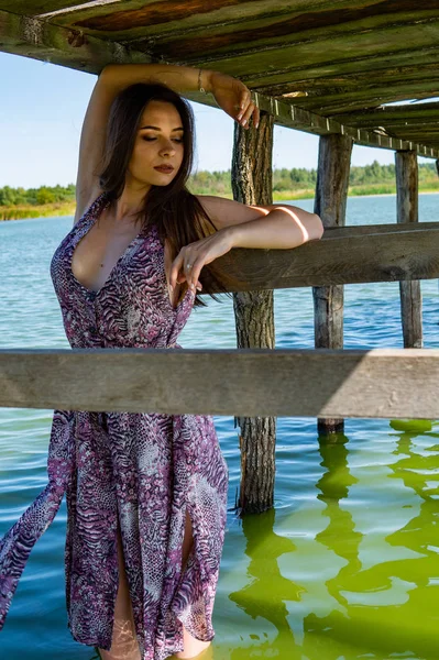 Dark-haired woman posing at the wooden pier at the lake. Woman in summer outfit with her back turned to the wooden footbridge of Lake Neusiedl. Sun reflections at the boathouse in the background. — Stock Photo, Image