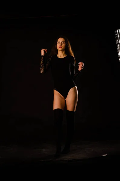 Jonge vrouw in een zwart bodysuit poseren op een donkere achtergrond, zwart haar en een gespierde sportfiguur. — Stockfoto