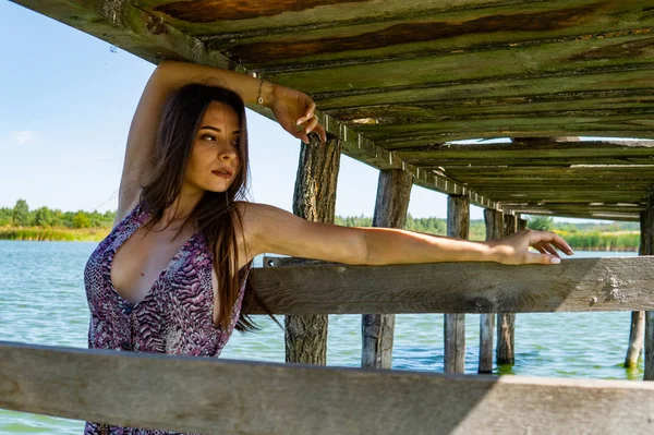 Dark-haired woman posing at the wooden pier at the lake. Woman in summer outfit with her back turned to the wooden footbridge of Lake Neusiedl. Sun reflections at the boathouse in the background. — Stock Photo, Image
