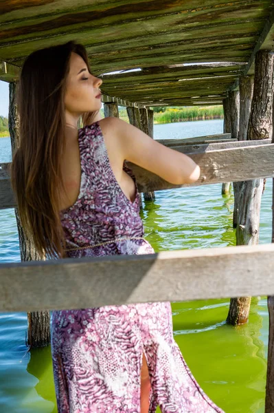 Donkerharige vrouw poseert bij de houten pier bij het meer. Vrouw in zomerkleding met haar rug naar de houten voetgangersbrug van het Neusiedlmeer. Zon reflecties bij het boothuis op de achtergrond. — Stockfoto