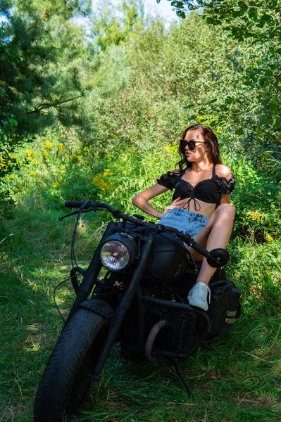 Młoda seksowna kobieta z długimi falowanymi włosami w skórzanych ubraniach i okularach przeciwsłonecznych, pozująca w pobliżu motocykla. Czarno-białe zdjęcie. — Zdjęcie stockowe