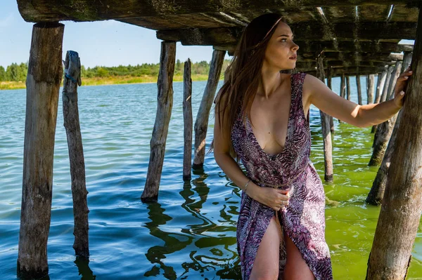 Femme aux cheveux bruns posant à la jetée en bois au lac. Femme en tenue d'été le dos tourné vers la passerelle en bois du lac Neusiedl. Réflexions du soleil au hangar à bateaux en arrière-plan. — Photo