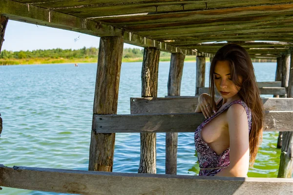 Dunkelhaarige Frau posiert am Holzsteg am See. Frau im Sommeroutfit mit dem Rücken zur hölzernen Fußgängerbrücke des Neusiedler Sees. Sonnenreflexe am Bootshaus im Hintergrund. — Stockfoto
