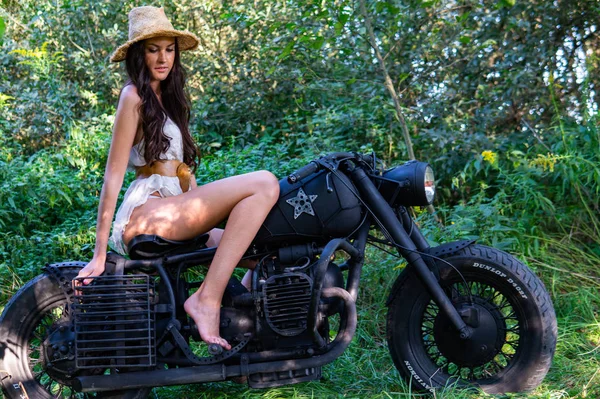 Jovem mulher sexy com cabelos longos ondulados em roupas de couro e óculos de sol posando perto de uma motocicleta. Foto em preto e branco . — Fotografia de Stock