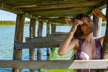 Gölün oradaki tahta iskelede poz veren siyah saçlı kadın. Sırtı Neusiedl Gölü 'nün tahta yaya köprüsüne dönmüş bir kadın. Arka plandaki kayıkhanede güneş yansımaları.