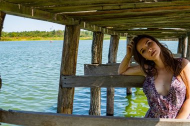 Gölün oradaki tahta iskelede poz veren siyah saçlı kadın. Sırtı Neusiedl Gölü 'nün tahta yaya köprüsüne dönmüş bir kadın. Arka plandaki kayıkhanede güneş yansımaları.