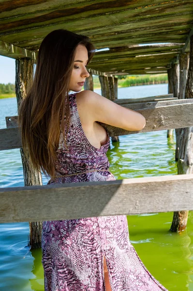 Tmavovlasá žena pózující u dřevěného mola u jezera. Žena v letních šatech se otočila zády k dřevěnému lávkovému můstku Neziderského jezera. Sluneční odrazy v loděnici v pozadí. — Stock fotografie