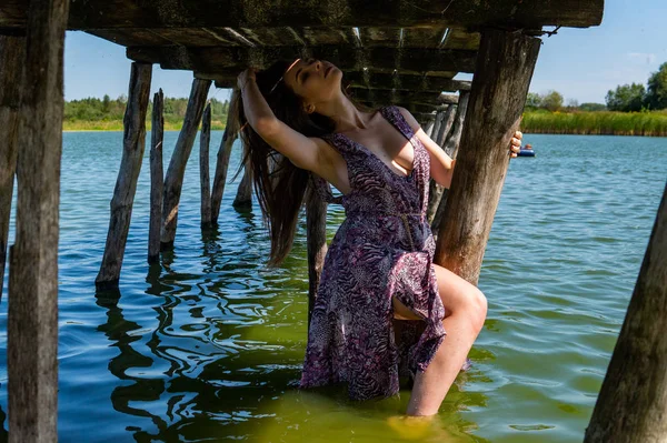Mulher de cabelos escuros posando no cais de madeira no lago. Mulher em roupa de verão com as costas viradas para a passarela de madeira do Lago Neusiedl. Reflexões do sol na casa do barco no fundo . — Fotografia de Stock