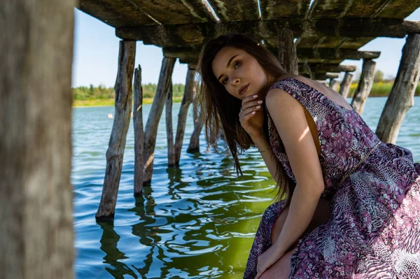 Mulher de cabelos escuros posando no cais de madeira no lago. Mulher em roupa de verão com as costas viradas para a passarela de madeira do Lago Neusiedl. Reflexões do sol na casa do barco no fundo . — Fotografia de Stock