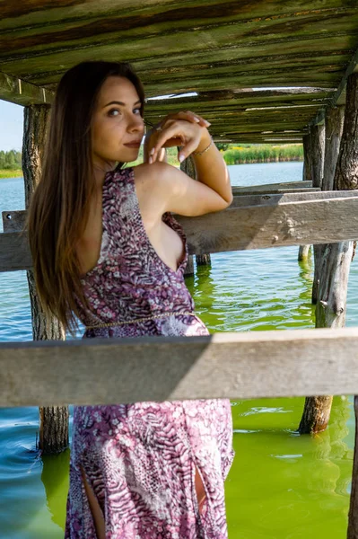 Ciemnowłosa kobieta pozująca na drewnianym pomoście nad jeziorem. Kobieta w letnim stroju z plecami odwróciła się do drewnianej kładki nad jeziorem Neusiedl. Odbicia słońca w hangarze na łodzi w tle. — Zdjęcie stockowe