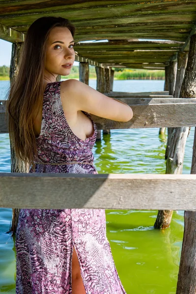 Donkerharige vrouw poseert bij de houten pier bij het meer. Vrouw in zomerkleding met haar rug naar de houten voetgangersbrug van het Neusiedlmeer. Zon reflecties bij het boothuis op de achtergrond. — Stockfoto