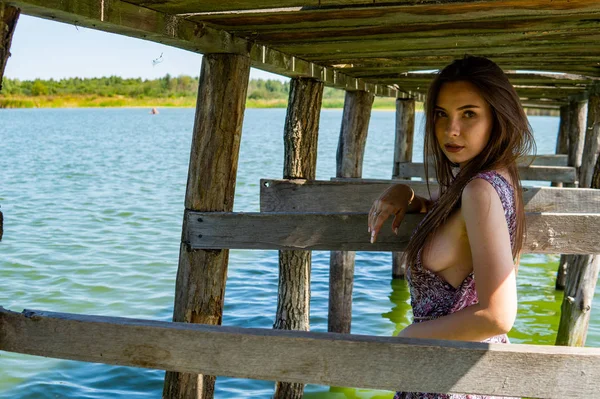 Donkerharige vrouw poseert bij de houten pier bij het meer. Vrouw in zomerkleding met haar rug naar de houten voetgangersbrug van het Neusiedlmeer. Zon reflecties bij het boothuis op de achtergrond. — Stockfoto