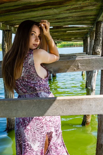 Donna dai capelli scuri posa al molo di legno al lago. Donna in abito estivo con la schiena rivolta verso la passerella in legno del lago Neusiedl. Riflessioni del sole alla rimessa sullo sfondo. — Foto Stock