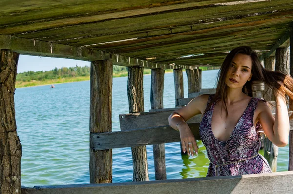 Mulher de cabelos escuros posando no cais de madeira no lago. Mulher em roupa de verão com as costas viradas para a passarela de madeira do Lago Neusiedl. Reflexões do sol na casa do barco no fundo . — Fotografia de Stock