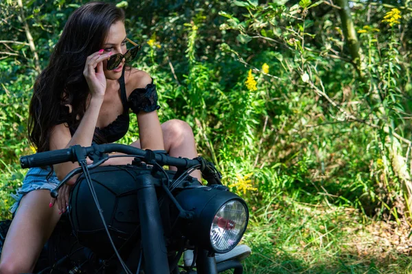 Jonge sexy vrouw met lang golvend haar in lederen kleding en zonnebril poseren in de buurt van een motorfiets. Zwart-wit foto. — Stockfoto