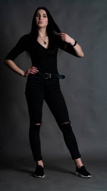 Yüksek tahta koltukta bacak bacak üstüne atmış esmer bir kadının portresi. Geniş paçalı pantolon giyen şık bir kız. Gri arkaplanda stüdyo çekimi.