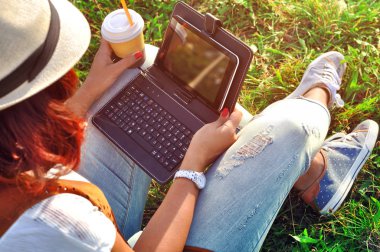 Kahve ve tablet ile bir fincan gevşeme. Kız ile laptop bir kahve. Kot pantolon, beyaz gömlek ve şapka giyen kadın, öğrenci çimlere oturup dizüstü kullanarak. Açık. Güneşli gün. 