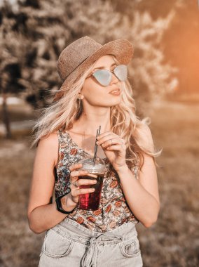Smiling caucasian woman in hat with coctails has fun outdoors at sunset.