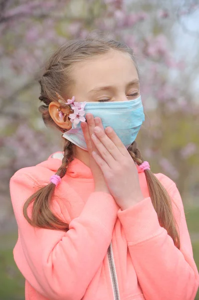 Cute girl in protective medicine mask with branche of blooming cherry. Quarantine. Coronavirus pandemic.