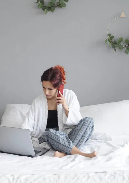 Upset woman looks at laptop and talks on the phone in bed at home.